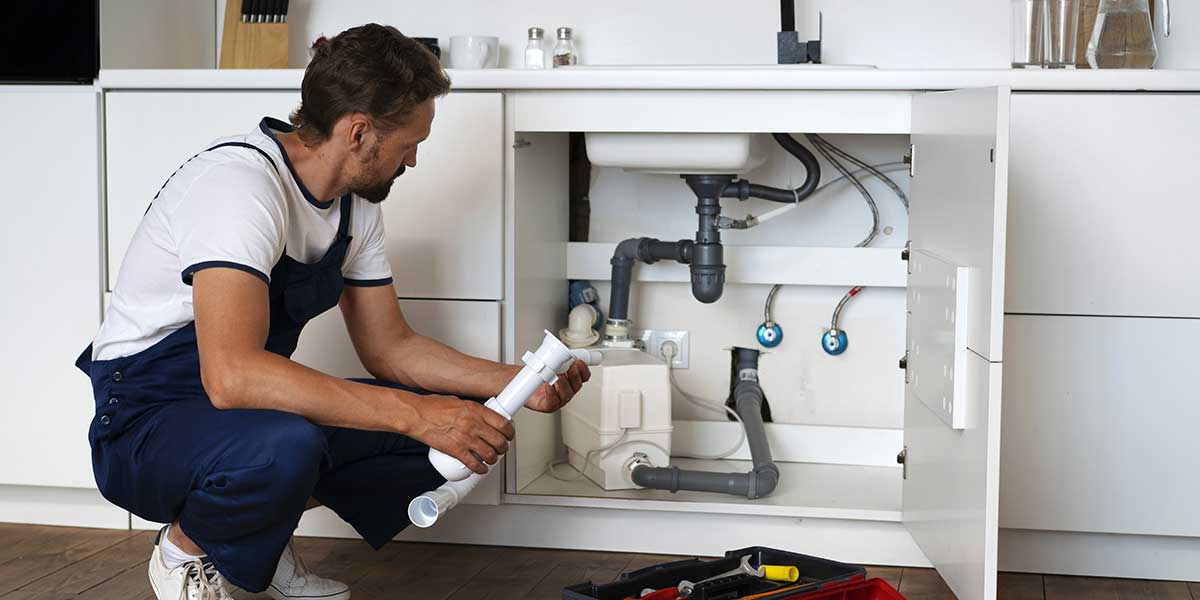 A plumber in overalls crouches before an open cabinet, examining a white PVC pipe section to repair kitchen sink plumbing.